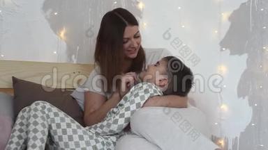 一位美丽的母亲和坐在床上的儿子玩，吻他有一个好心情。